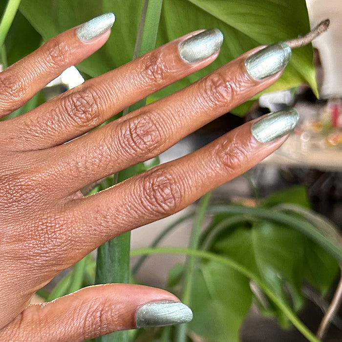 Hand with Jade Velvet cool green magnetic polish showing subtle shine against green leafy background for manicure look