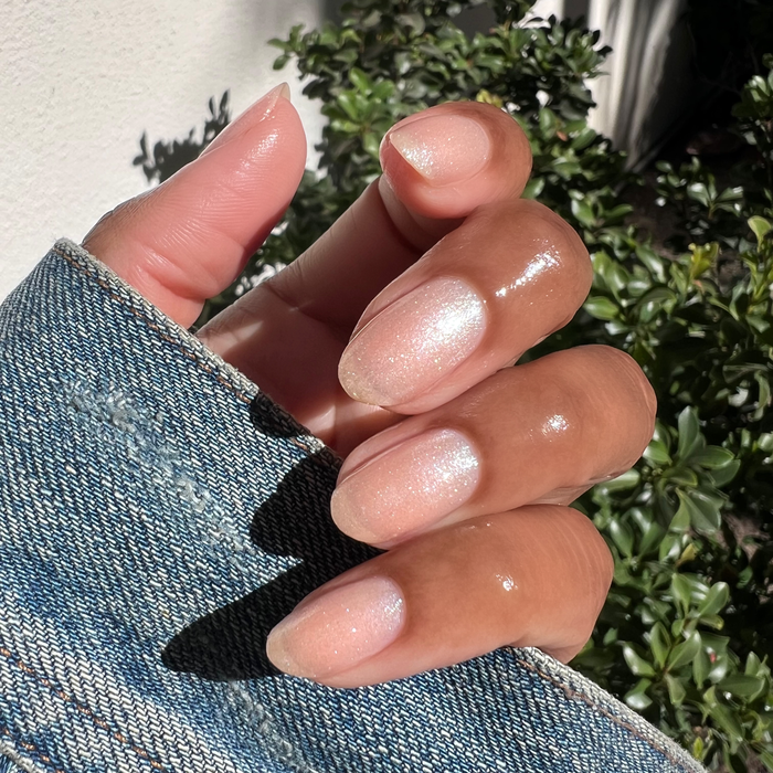 Hand with natural medium nails coated in warm shimmering Starfruit Shimmer Gloss polish outdoors with greenery background
