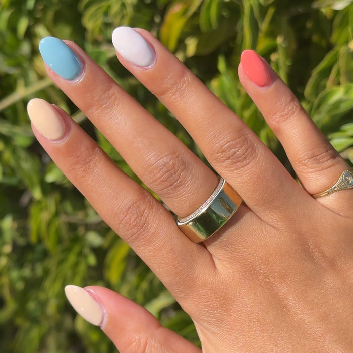 Hand with pastel blue, white, coral, and cream nails showcasing Sunset Season polished mani against green leaves background