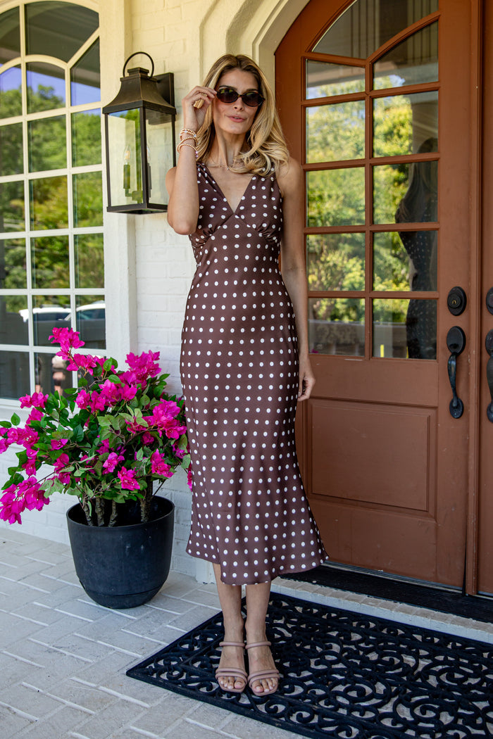 A woman wearing a brown and white polka dot midi dress stands on a front porch next to a potted plant with bright pink flowers.