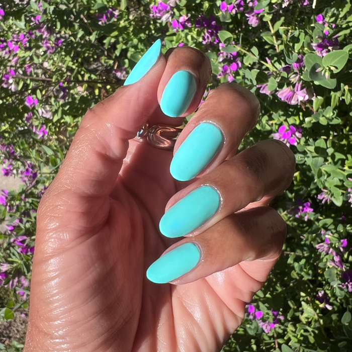 Hand with nails painted in bright blue-green All Manis on Deck gel polish showing smooth manicure outdoors with floral background