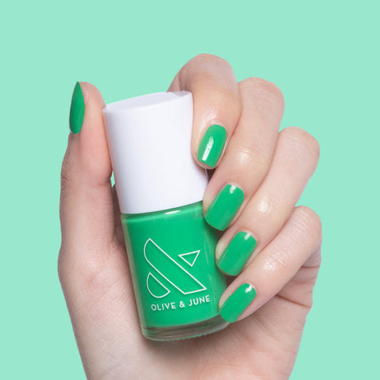 Hand holding Olive & June Bocce Ball nail polish bottle with matching bright green manicure on mint background