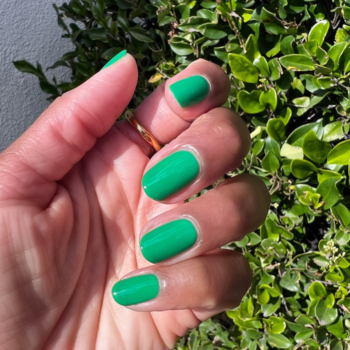 Hand with polished nails in vibrant Bocce Ball green shade against leafy outdoor background in natural light