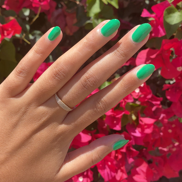 Hand with nails painted in vivid Bocce Ball green polish against bright pink flowers in natural sunlight