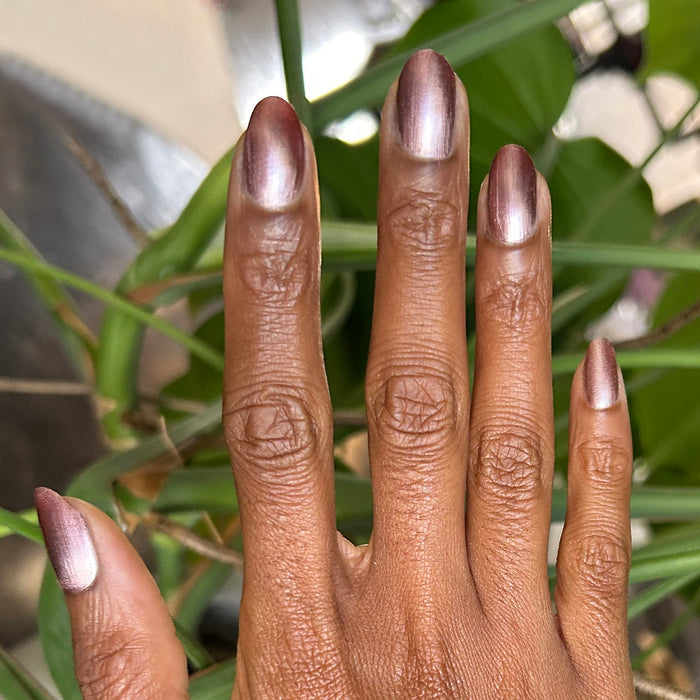Hand with Bronze Velvet nails in metallic neutral pink shade displayed against green leafy background for manicure inspiration