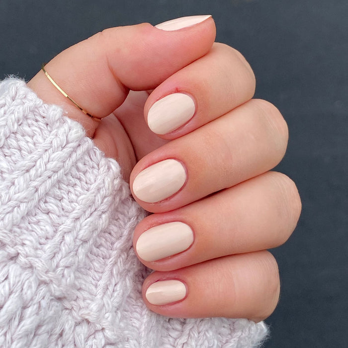 Hand with nails painted in Cable Knit cream peach gel polish wearing delicate gold rings and cozy cable knit sweater sleeve