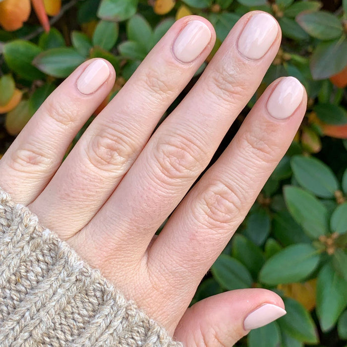 Hand with nails polished in Cable Knit soft cream peach gel polish wearing tan cable knit sweater sleeve outdoors