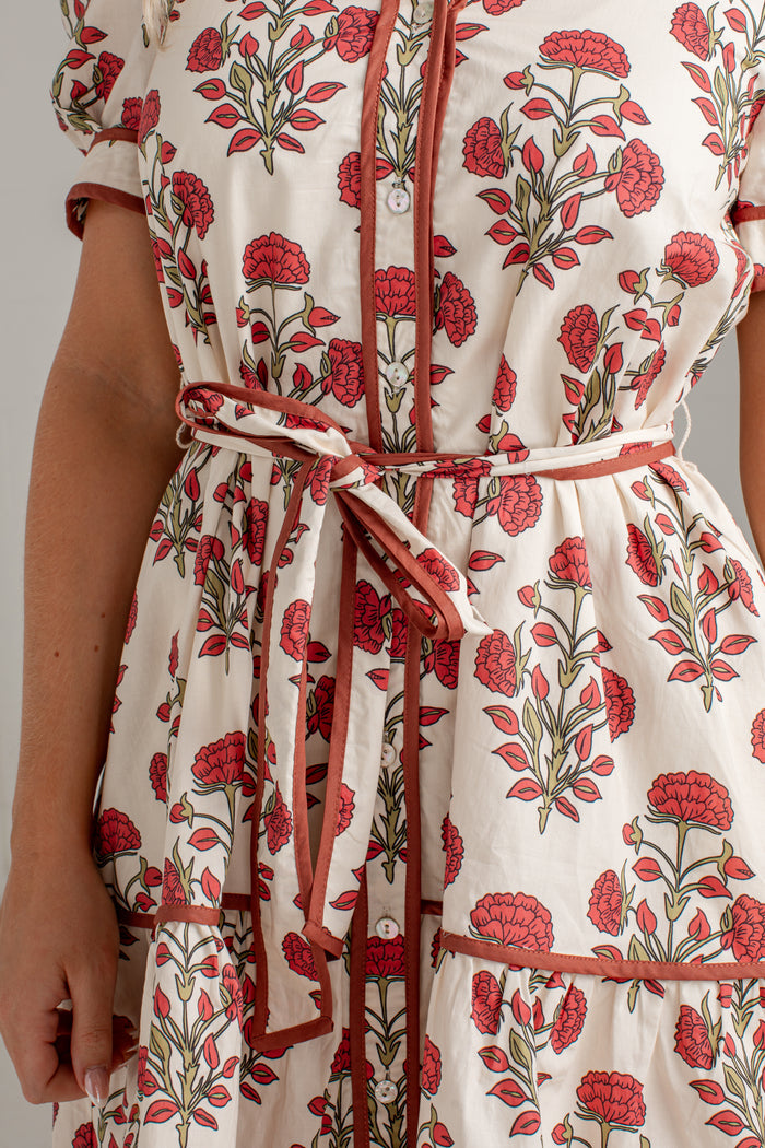 Close-up of a cream-colored dress with a red and green floral print, featuring a tie waist belt and red trim.