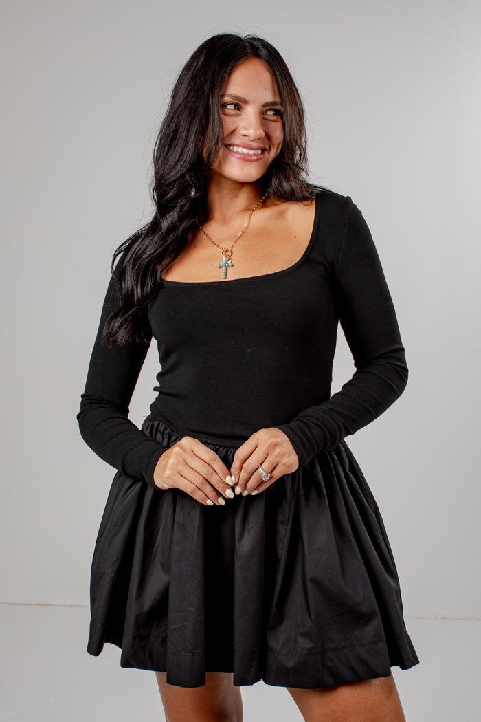 A woman modeling a black long-sleeve scoop-neck top with a matching black pleated mini skirt, photographed against a neutral light gray studio background.