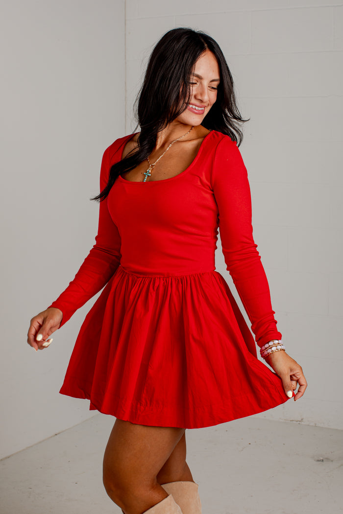 A woman wearing a bright red long-sleeve skater dress with a scoop neckline, standing against a plain light gray wall.