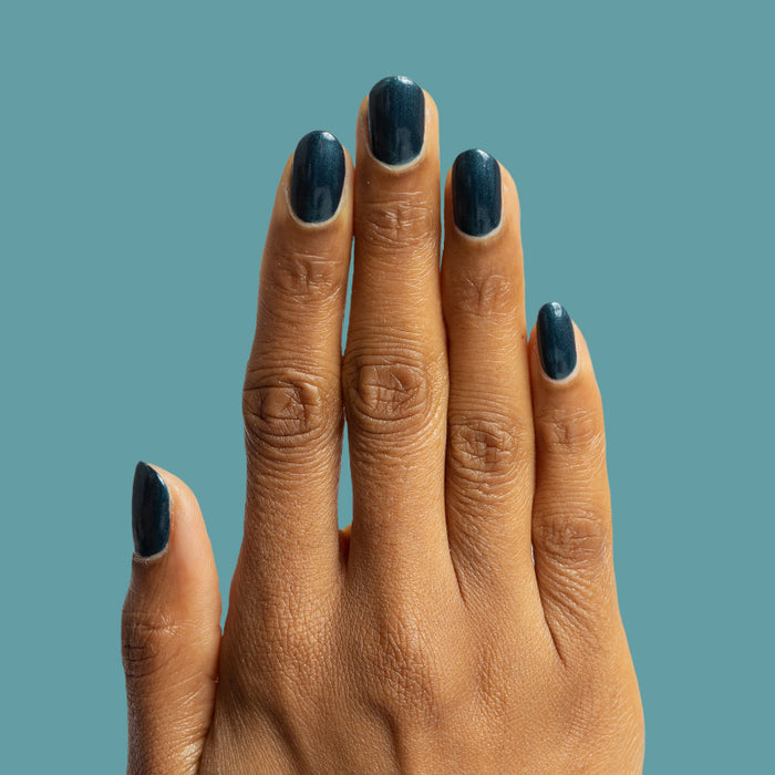 Hand with medium-length nails painted in deep Dancing Green polish against muted teal background for manicure display