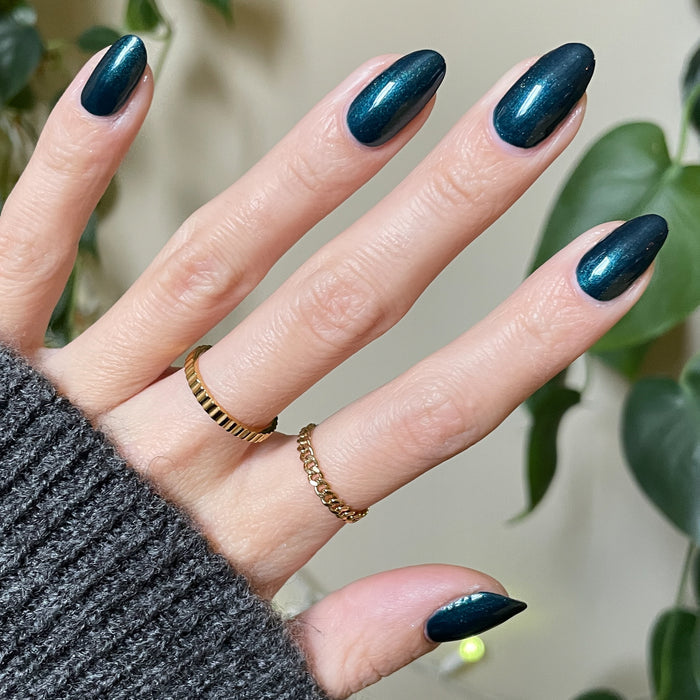 Hand with long almond-shaped nails painted in shimmering Dancing Green polish wearing gold rings against plant background