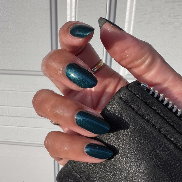 Hand with medium-length nails polished in shimmering Dancing Green holding black textured leather item in natural light