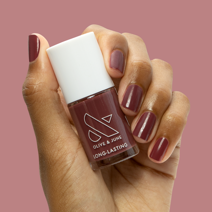 Hand with short nails painted in Don’t Spill on the White Couch berry red polish holding matching Olive and June bottle