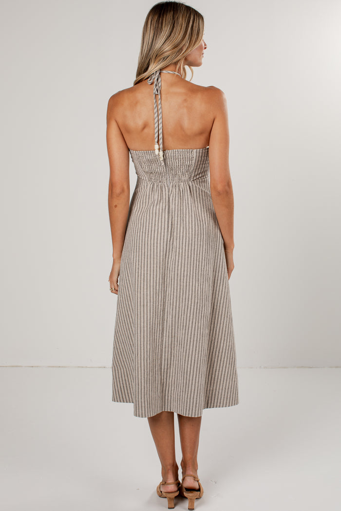 A woman viewed from the back wearing a sleeveless halter midi dress with thin vertical pinstripes in beige and white, paired with tan heeled sandals against a plain light gray studio background.