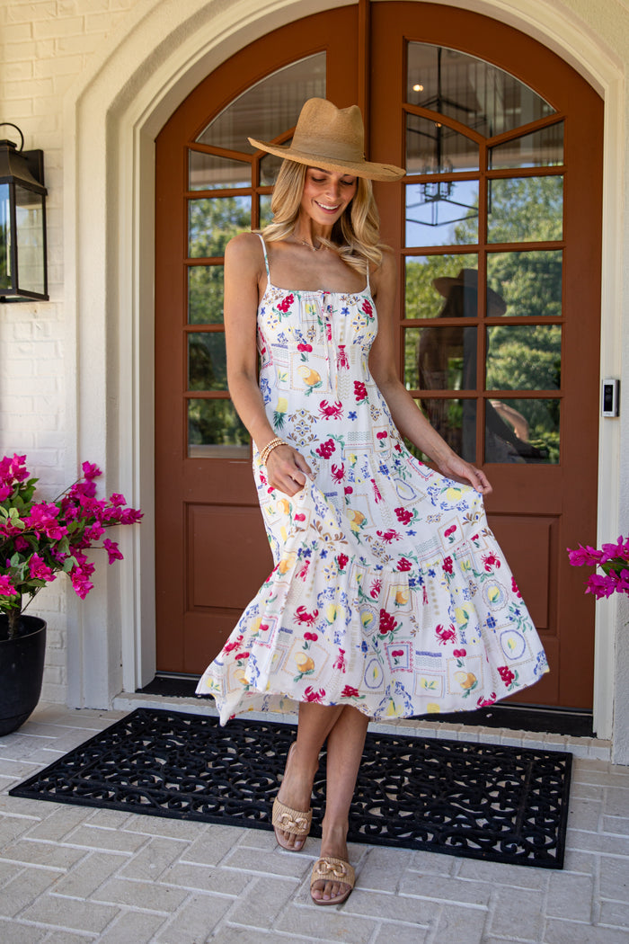 Woman in a floral dress and wide-brimmed hat standing in front of a house entrance.