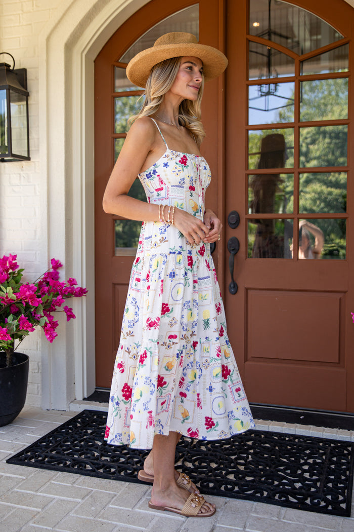 Woman in a floral dress and wide-brimmed hat standing in front of a house entrance.