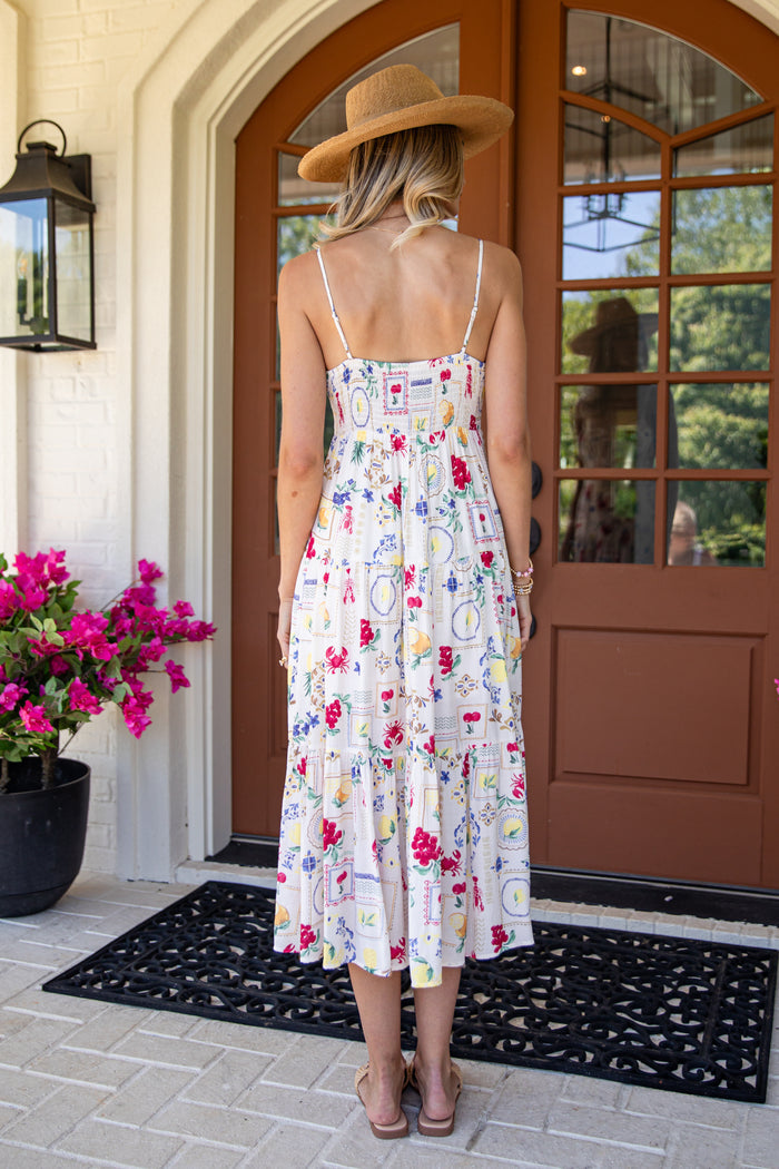 Woman in a floral dress and wide-brimmed hat standing in front of a house entrance.