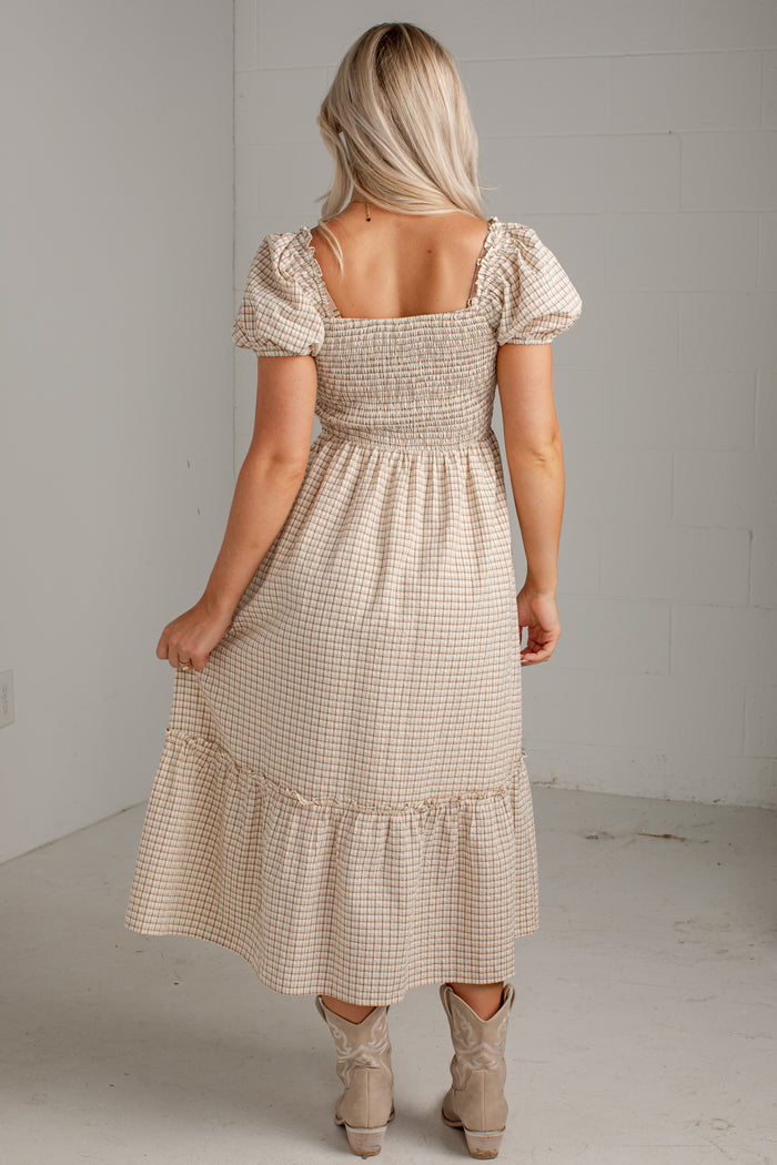 A woman modeling a beige and white gingham tiered midi dress with puff sleeves and a smocked bodice, paired with beige ankle boots, in a minimalist studio setting.