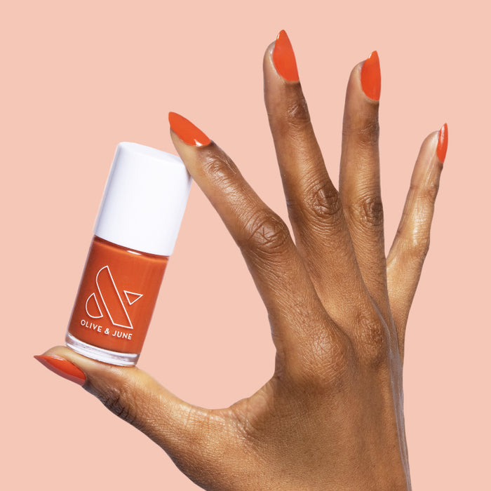 Hand with nails painted in burnt orange Flowerpot polish holding matching Olive and June bottle on peach background