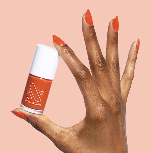 Hand with nails painted in burnt orange Flowerpot polish holding matching Olive and June bottle on peach background