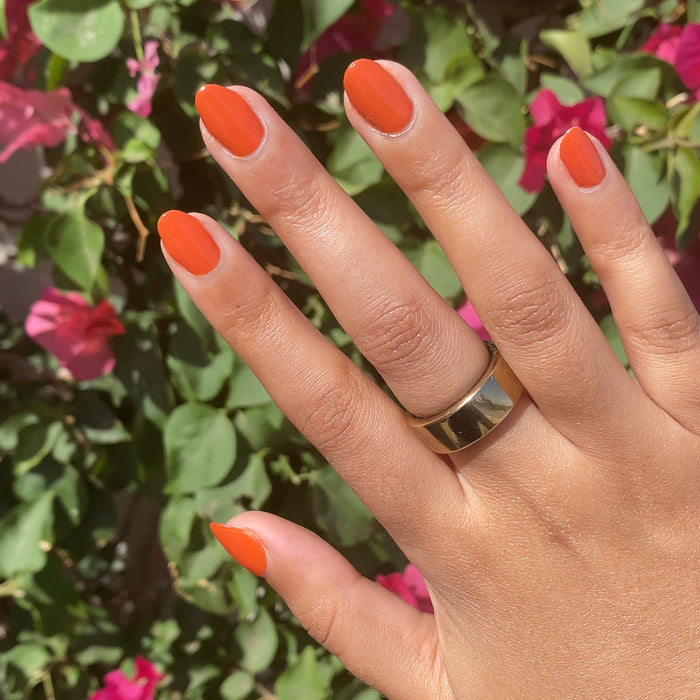 Hand with nails painted in bold Flowerpot burnt orange polish with gold ring and pink flowers in the background