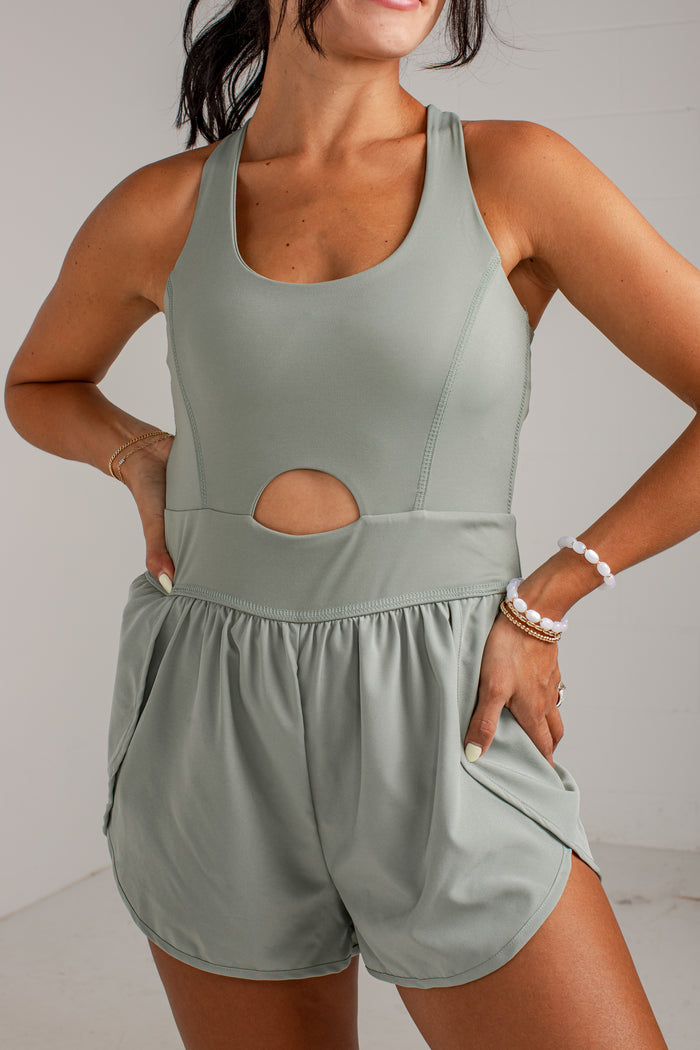 A woman wearing a sage green sleeveless romper with a scoop neckline, a side cutout, and matching shorts with side pockets, posed against a light gray wall.