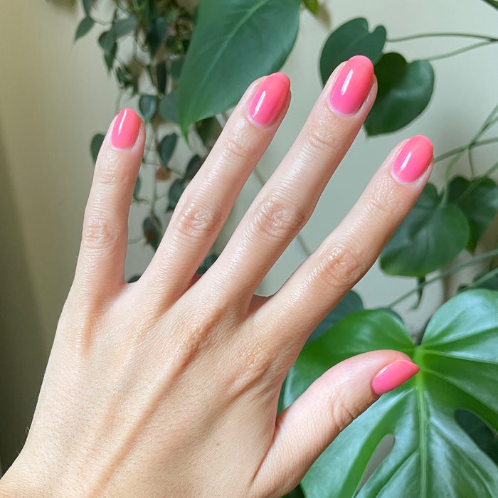 Hand with natural nails coated in bright pink Guava Gloss quick dry polish against a green leafy background