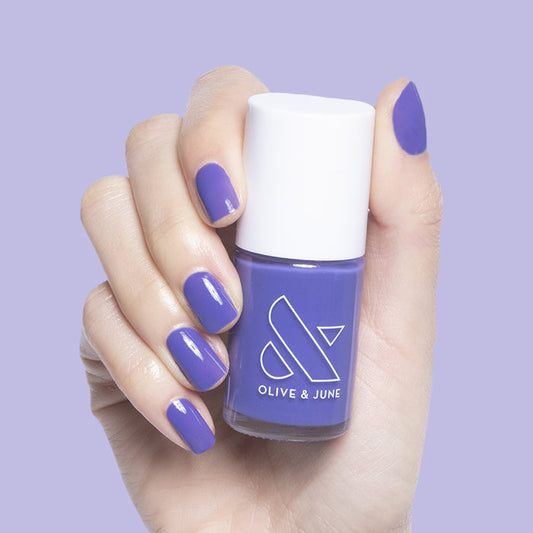 Hand with nails painted in deep purple House of O&J polish holding Olive and June bottle on lavender background