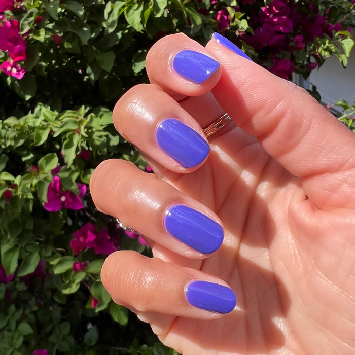 Hand with nails polished in vibrant royal purple House of O&J nail polish against green leaves and magenta flowers background