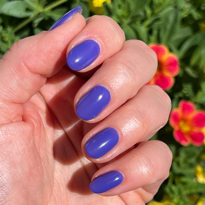 Hand with nails painted in vibrant glossy House of O&J polish shown outdoors with colorful flowers in background