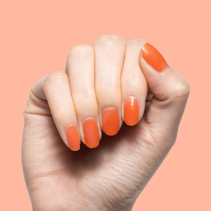 Hand with nails painted in I’ll Take Another Spritz showing a smooth red-orange manicure against peach background
