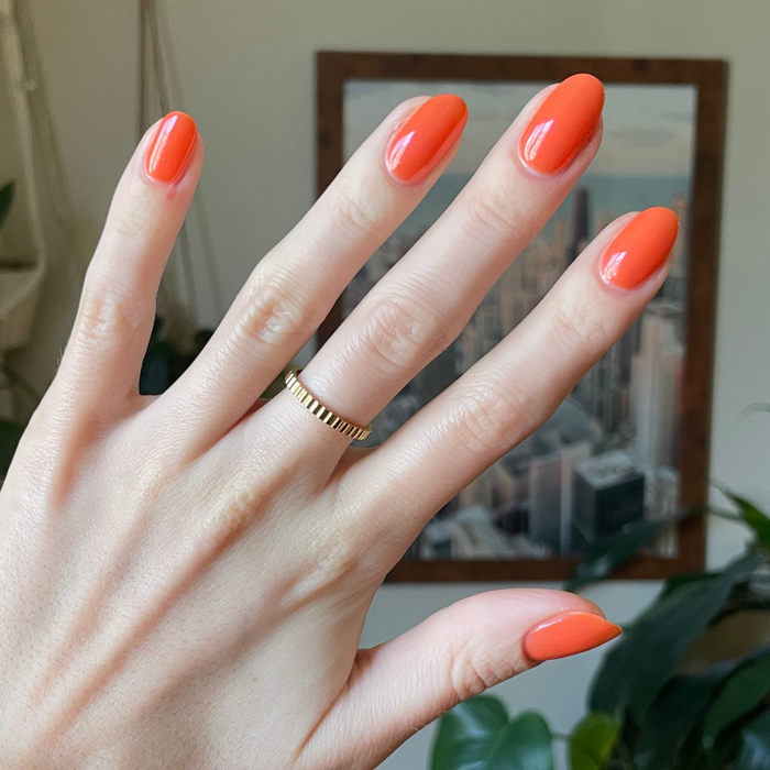Hand with almond-shaped nails in glossy I’ll Take Another Spritz gel polish showing vibrant red-orange manicure at home