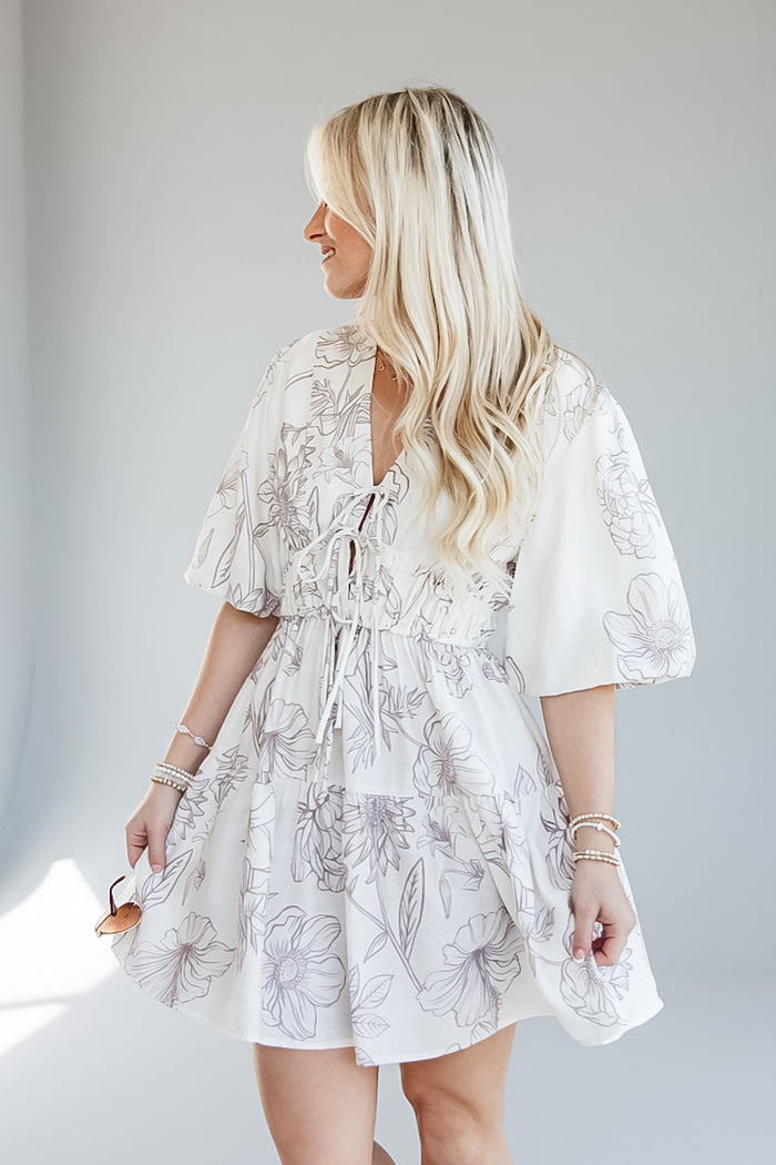 A woman wearing a white tiered mini dress with a gray floral line print, shown from the back-three-quarter view against a light gray studio background.