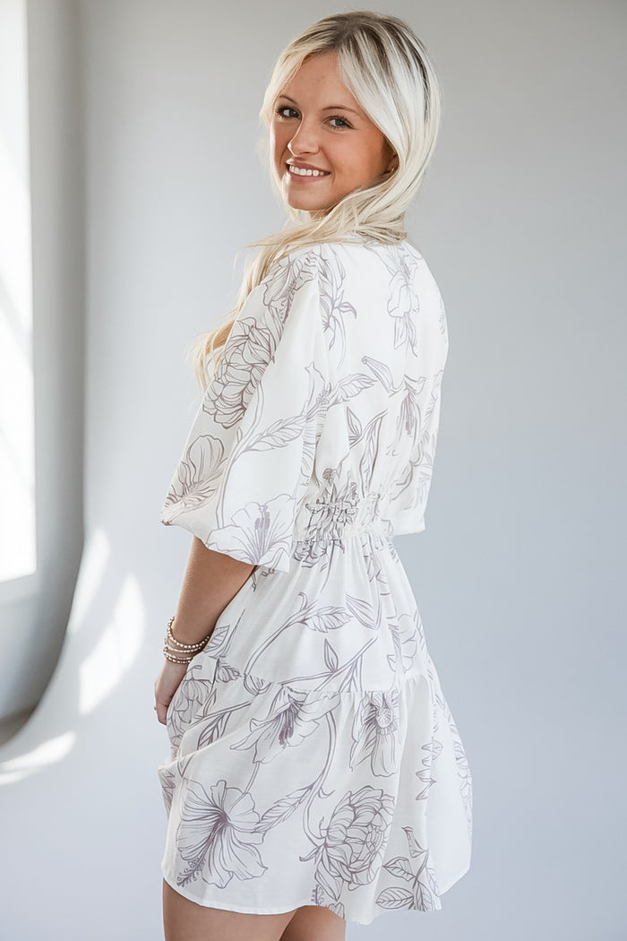 A woman wearing a white short-sleeve dress with a gray floral line-art print, photographed in a bright indoor setting.