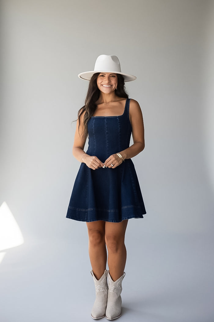 A woman modeling a dark blue denim dress with a white wide-brim hat and white cowboy boots, standing against a light gray studio background.