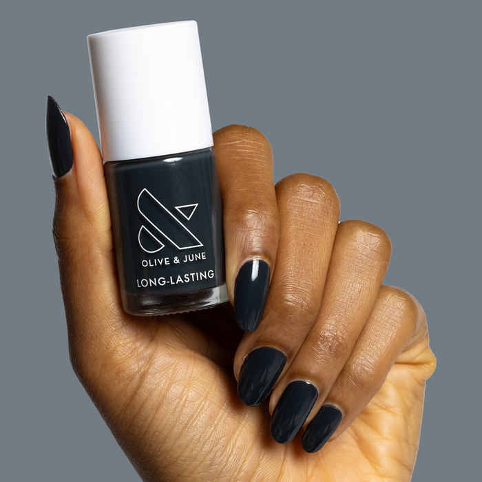 Hand holding Olive and June Last Minute Shopping polish bottle with nails painted deep charcoal gray manicure.
