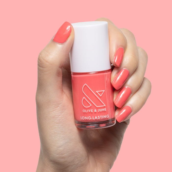Hand with nails painted in Maine Character Energy coral red gel polish holding an Olive and June long-lasting polish bottle