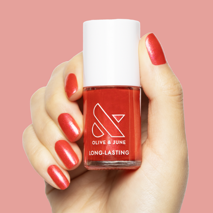 Hand holding Rodeo Queen polish bottle showing bright pinky-red nails with silver shimmer for long-lasting mani