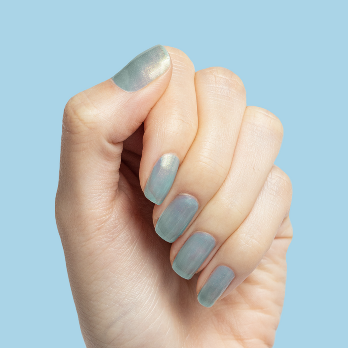 Hand with sheer blue iridescent nails painted with Sequin Everything polish against a soft blue background
