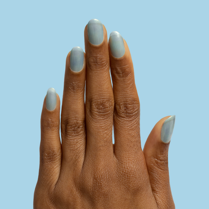Hand with medium-length nails painted with Sequin Everything sheer blue iridescent polish against light blue background