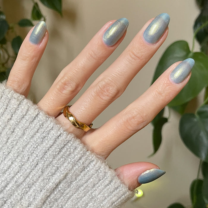Hand wearing gold band with nails painted in shimmering blue-green shades using Sequin Everything polish against neutral background