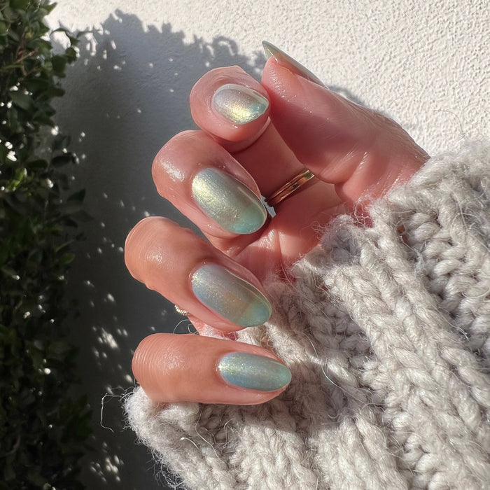 Hand with short rounded nails painted with Sequin Everything sheer blue iridescent polish in natural light outdoors