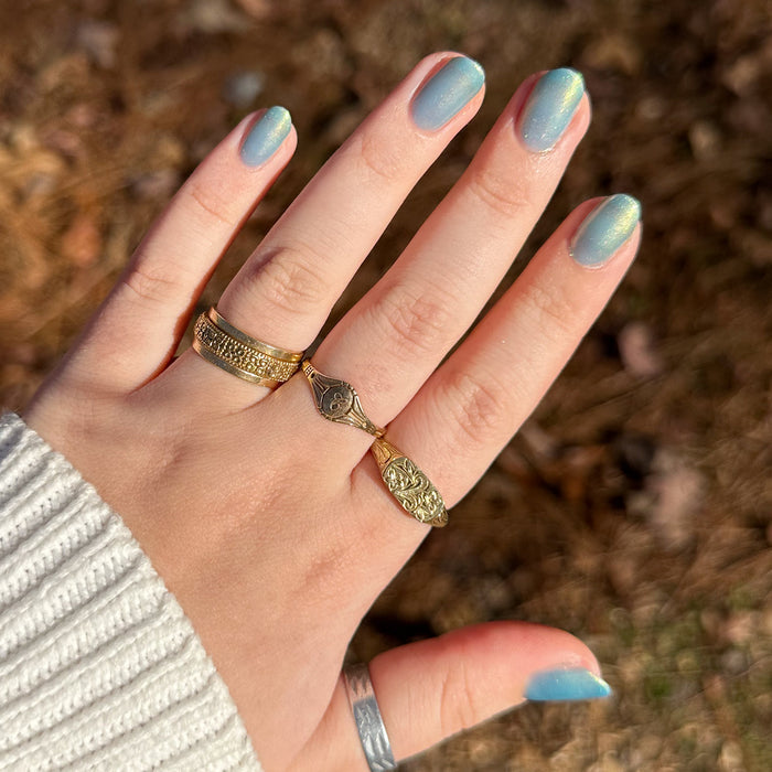 Hand with short nails painted in sheer blue iridescent Sequin Everything polish wearing gold rings outdoors