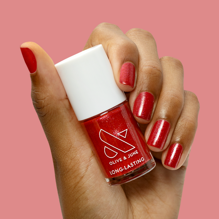 Hand holding Splash of Cran nail polish bottle with shimmery bubbly pinky-red mani on short nails against pink background