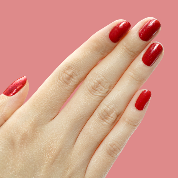 Hand with short nails painted in shimmery berry red gel polish Splash of Cran against a pink background