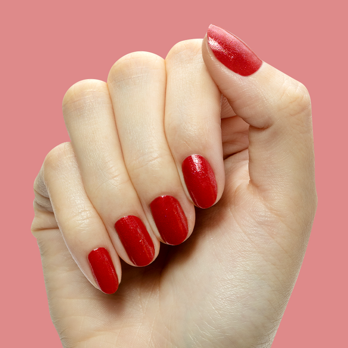 Hand with short nails painted in shimmery bubbly red gel polish Splash of Cran against a coral background