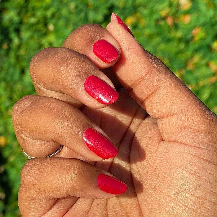 Hand with short nails painted in shimmery pinky-red Splash of Cran gel polish against green foliage background