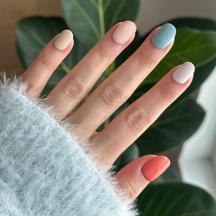 Hand with pastel and coral nail polish manicure in Sunset Season set shown against green leafy background