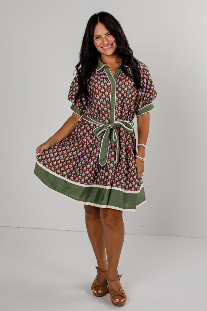 A woman modeling a short-sleeve button-front dress with an allover brown and white floral pattern, olive green trim, and a matching green tie belt, photographed against a plain light gray studio background.